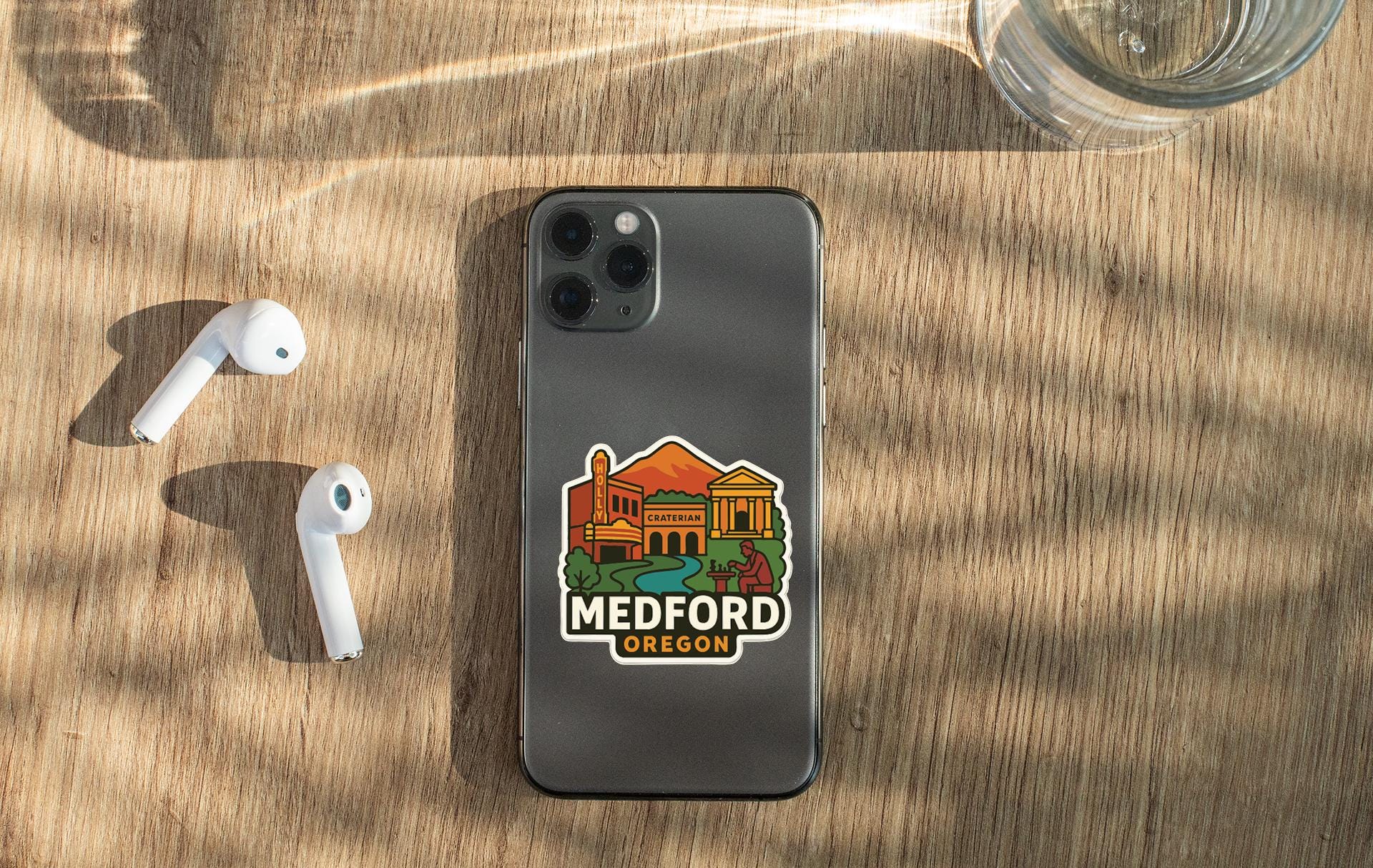 a smartphone with a sticker of Medford, Oregon, placed on a wooden surface alongside a pair of white earbuds and a glass of water.