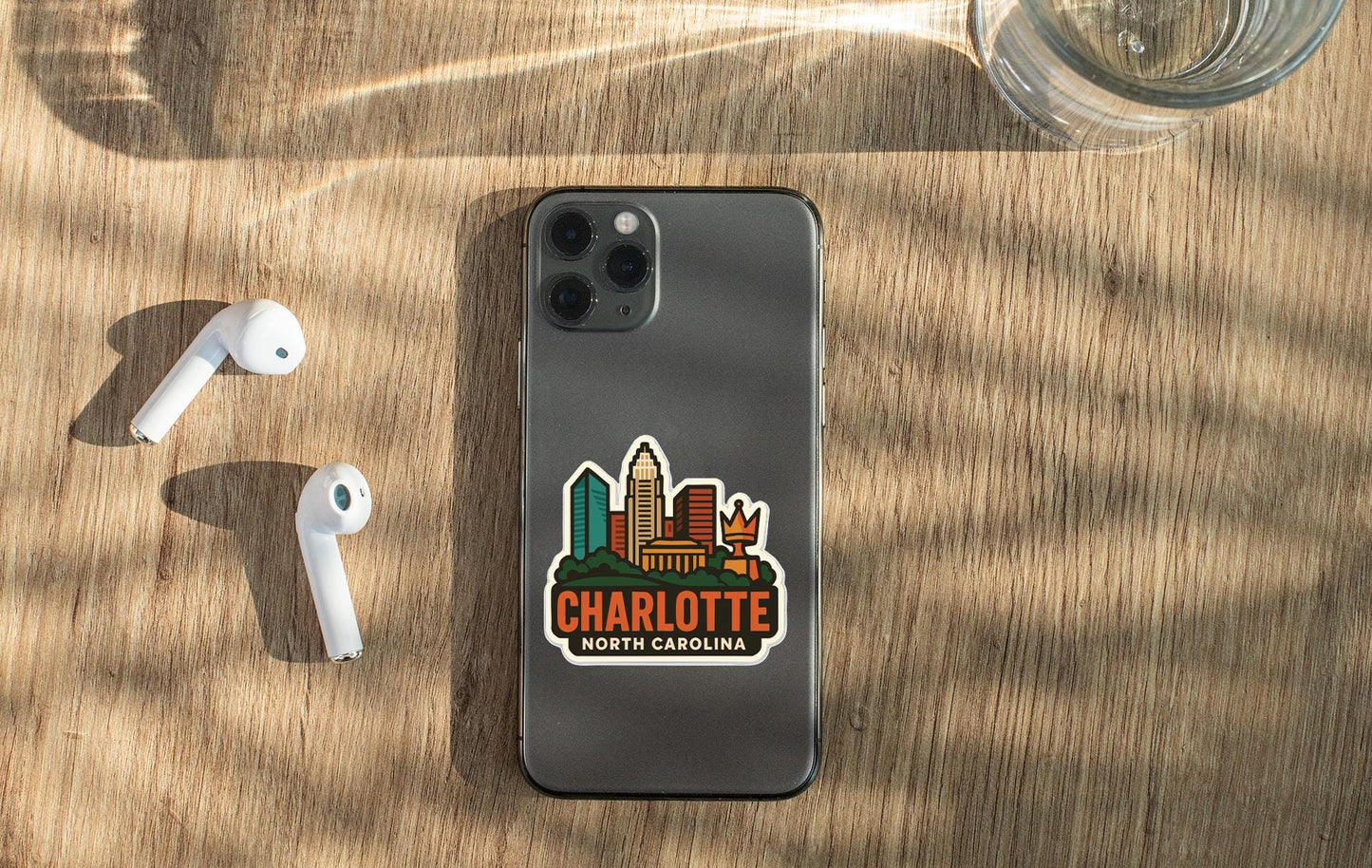 a smartphone with a sticker of the Charlotte, North Carolina skyline, along with a pair of white earbuds and a glass of water on a wooden surface.