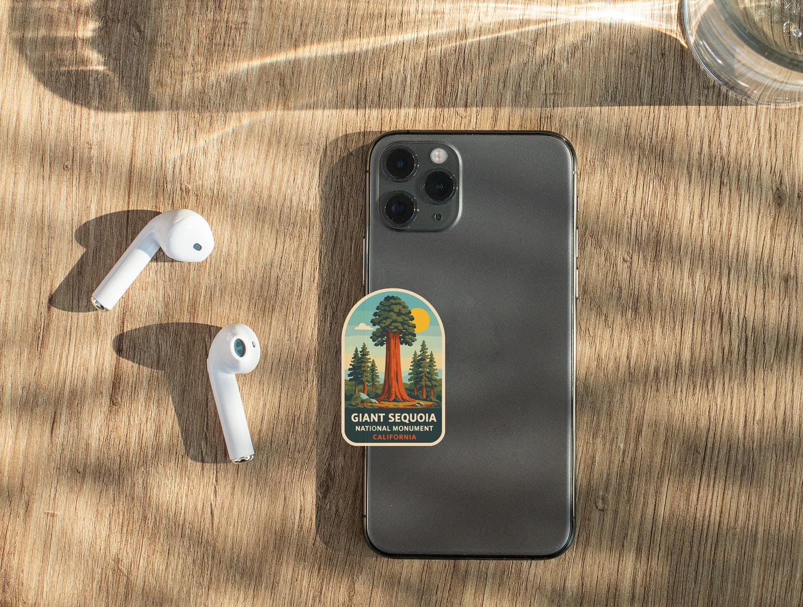 a smartphone with a sticker of Giant Sequoia National Park on it, along with a pair of white earbuds and a glass of water on a wooden surface.