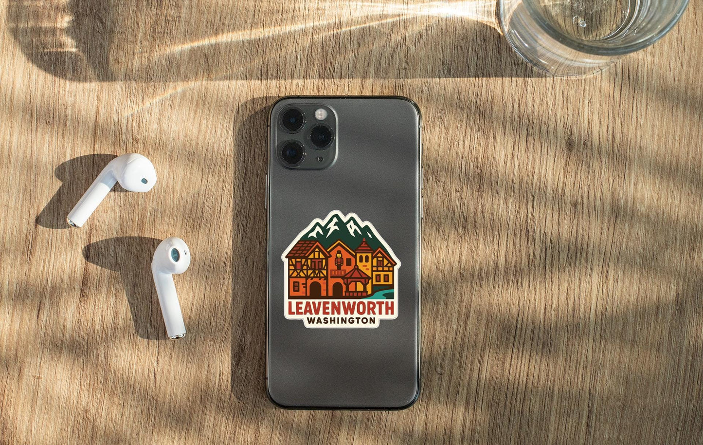 a smartphone with a sticker of a mountain town on it, placed on a wooden surface alongside a pair of white earbuds and a glass of water.