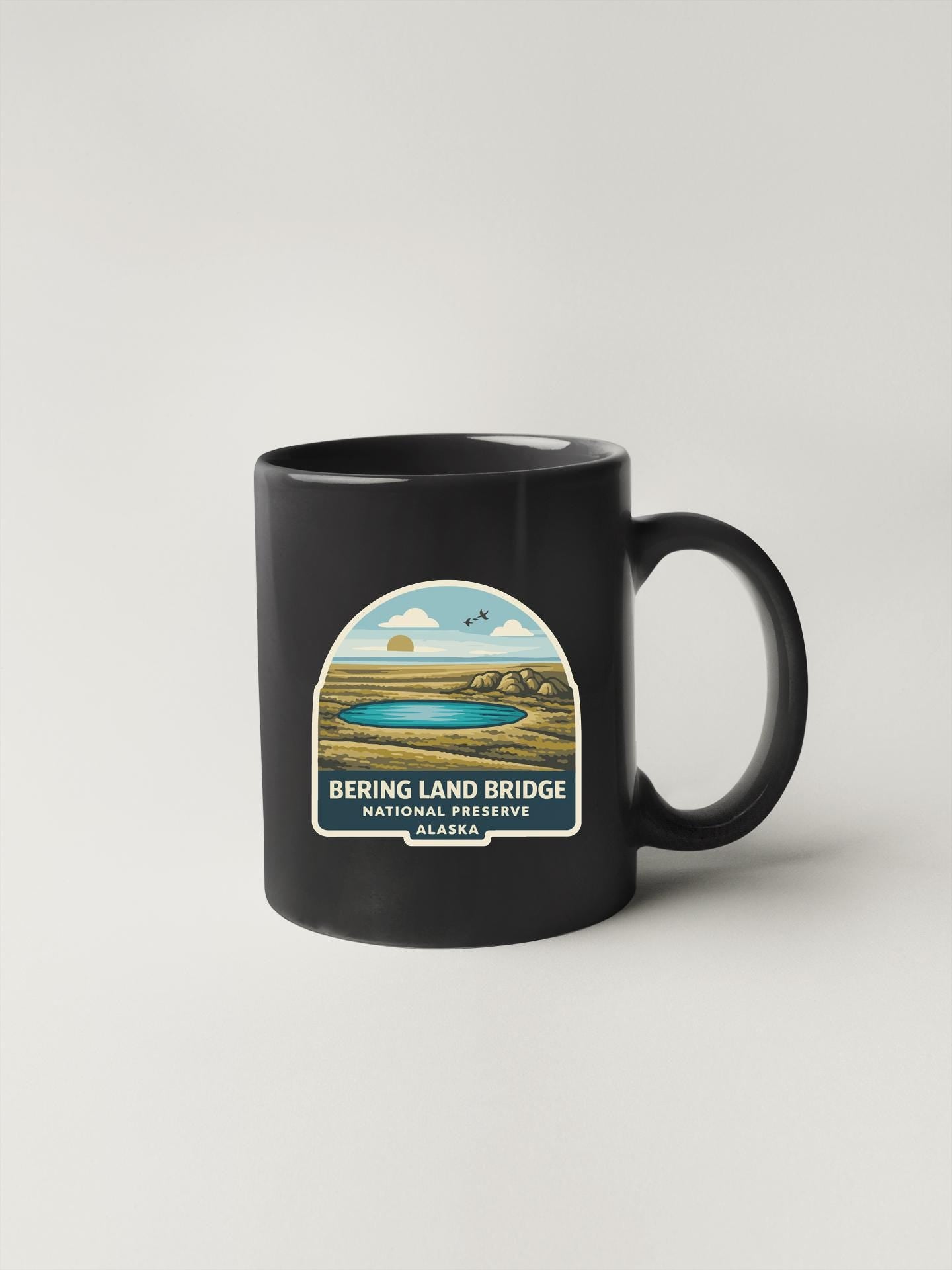 A black coffee mug with a label that says "BERING LAND BRIDGE NATIONAL PRESERVE WALKS" and features an illustration of a lake and mountains.