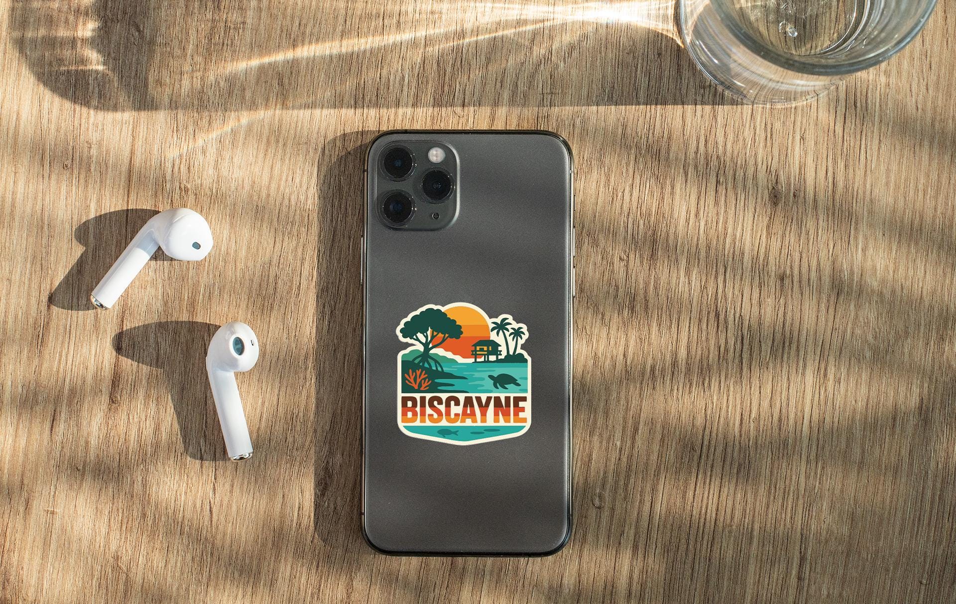 a smartphone with a sticker on the back, placed on a wooden surface alongside a pair of white earbuds and a glass of water.