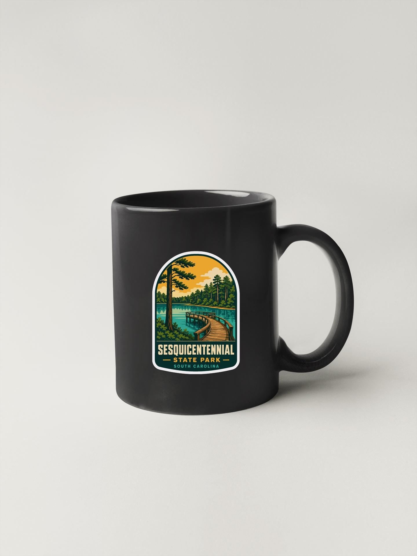 A black coffee mug with a graphic design depicting a scenic landscape, including a lake, trees, and a path.