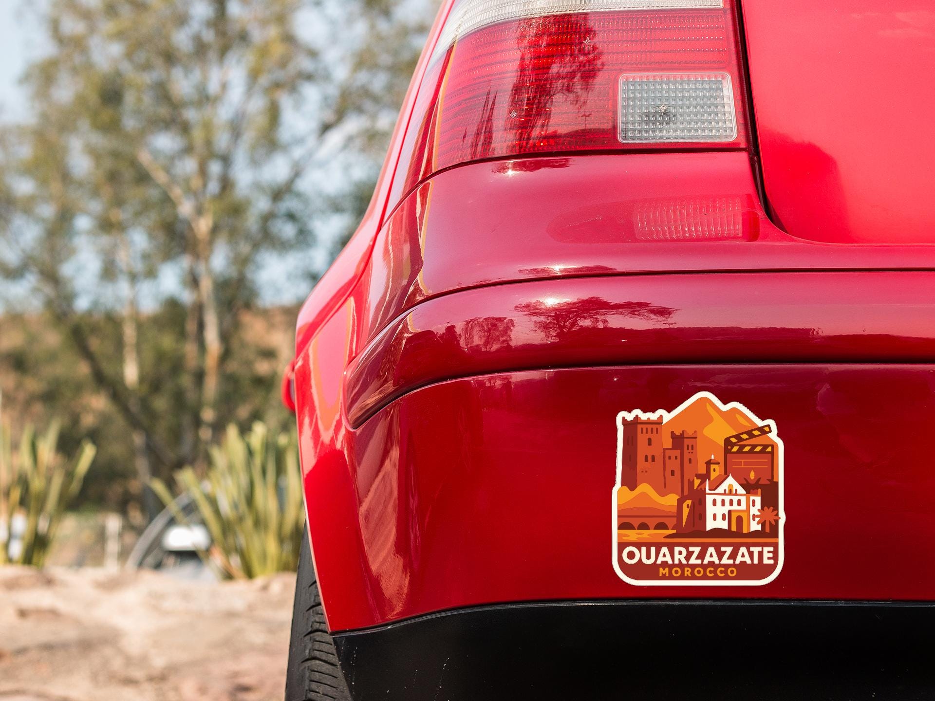 the rear end of a red car with a sticker on the trunk that features an illustration of a desert landscape with buildings and mountains.