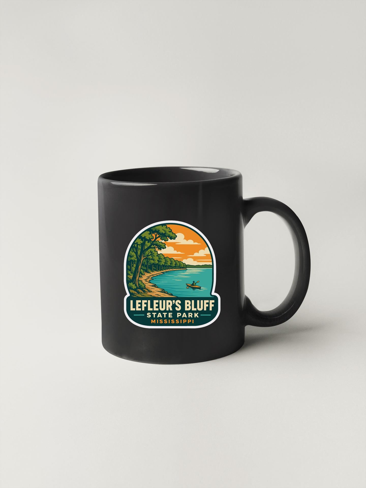 A black coffee mug with a graphic design depicting a scenic landscape, including a beach, mountains, and a lake.