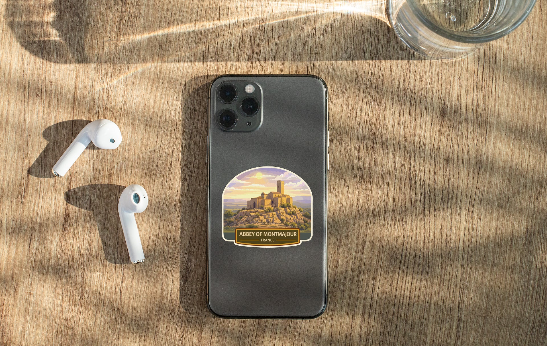 a smartphone with a sticker of a castle on it, placed on a wooden surface alongside a pair of white earbuds and a glass of water.