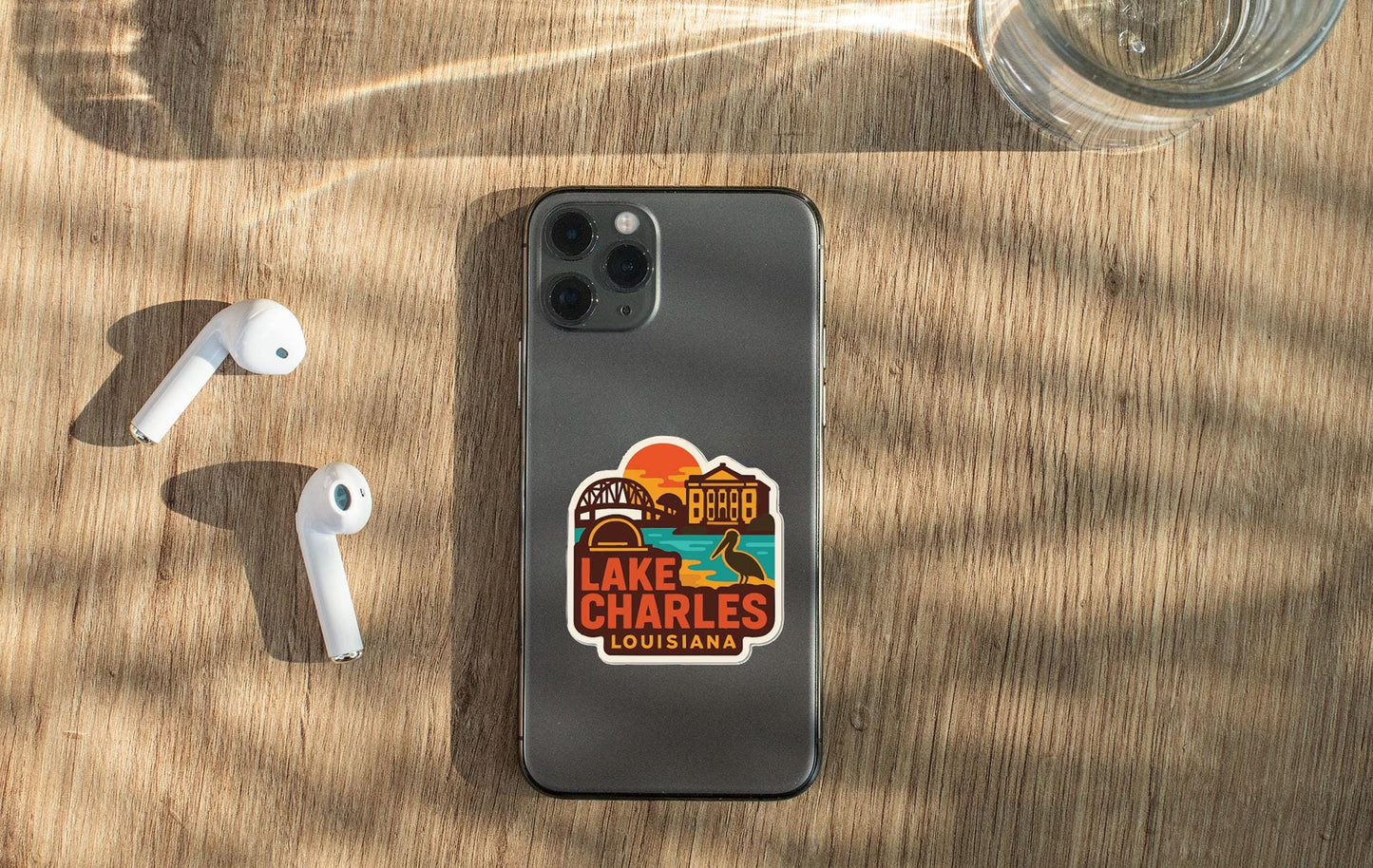 a smartphone with a sticker on it, placed on a wooden surface alongside a pair of white earbuds and a glass of water.