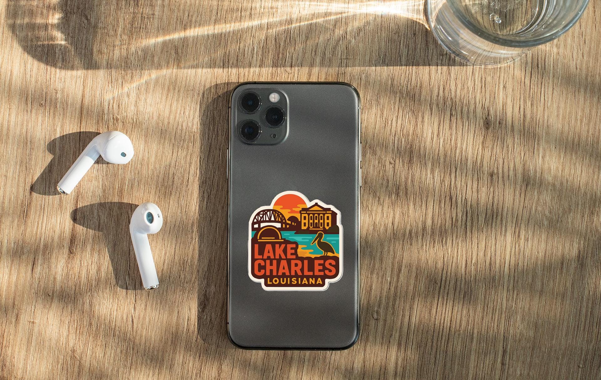 a smartphone with a sticker on it, placed on a wooden surface alongside a pair of white earbuds and a glass of water.