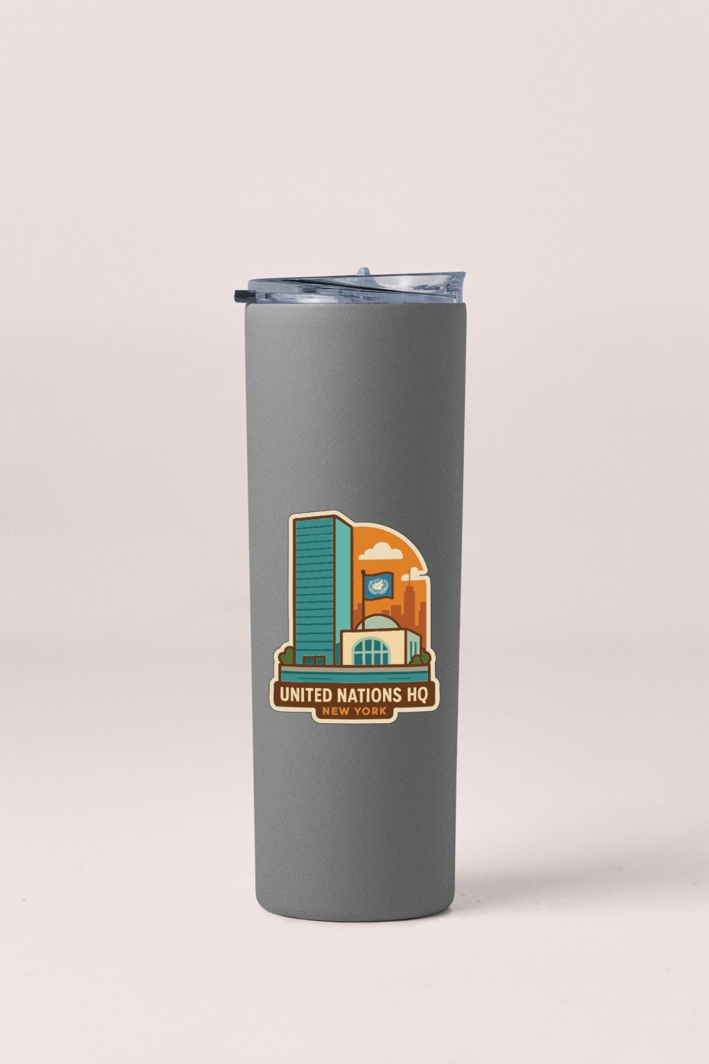 A gray insulated tumbler with a lid and a sticker featuring an illustration of a building and the text "UNITED NATIONS HQ NEW YORK".
