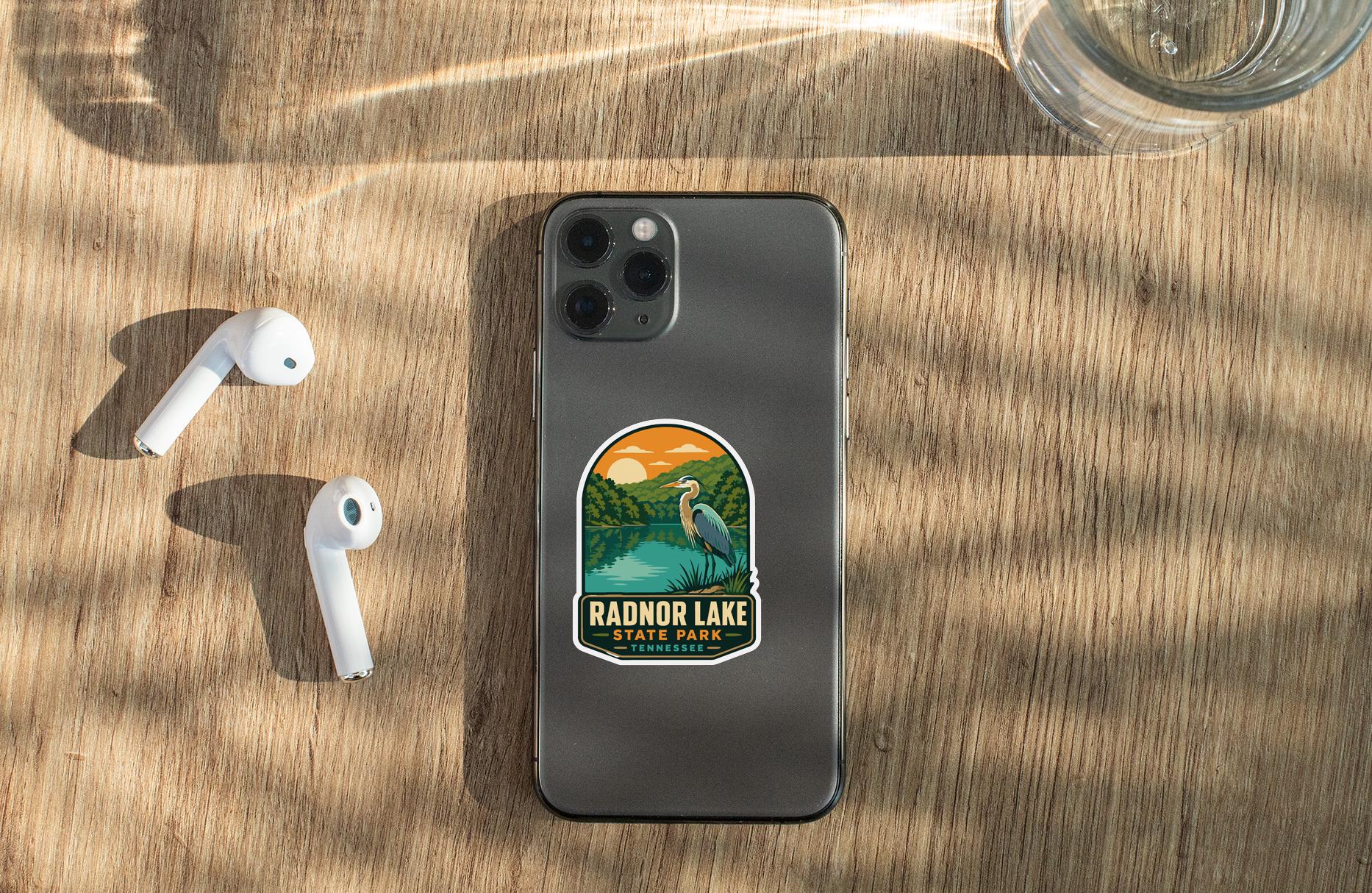 a smartphone with a sticker on the back, placed on a wooden surface alongside a pair of white earbuds and a glass of water.