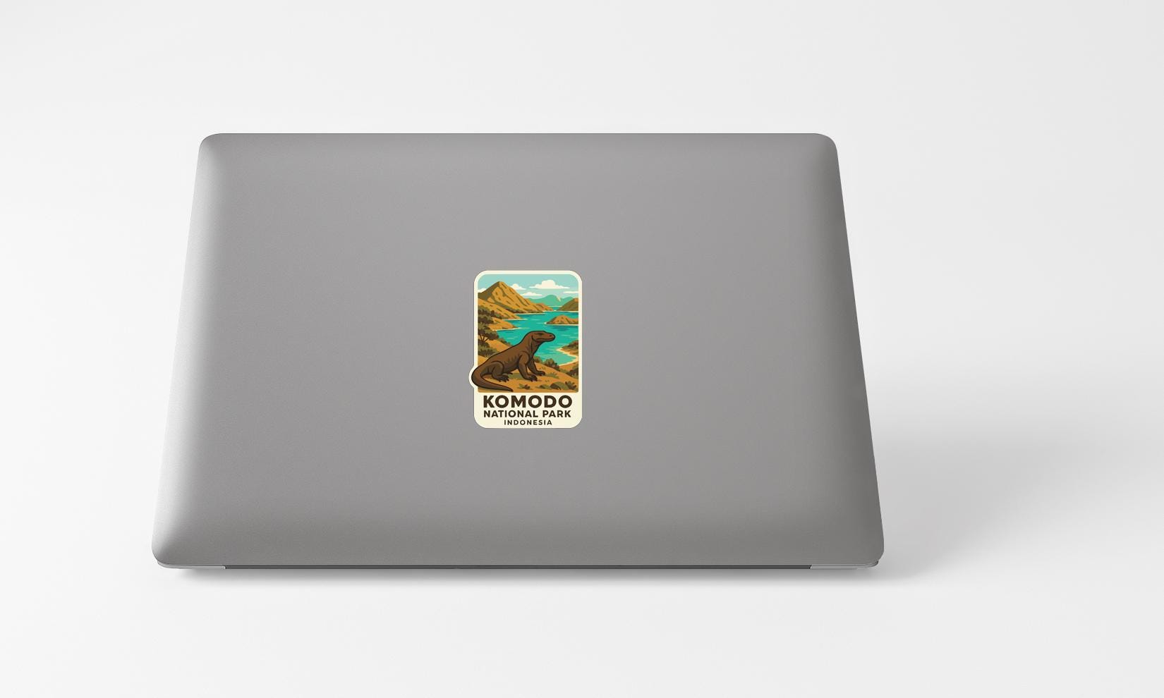 A sticker on the back of a laptop, depicting a scenic landscape with mountains, a lake, and a komodo dragon.