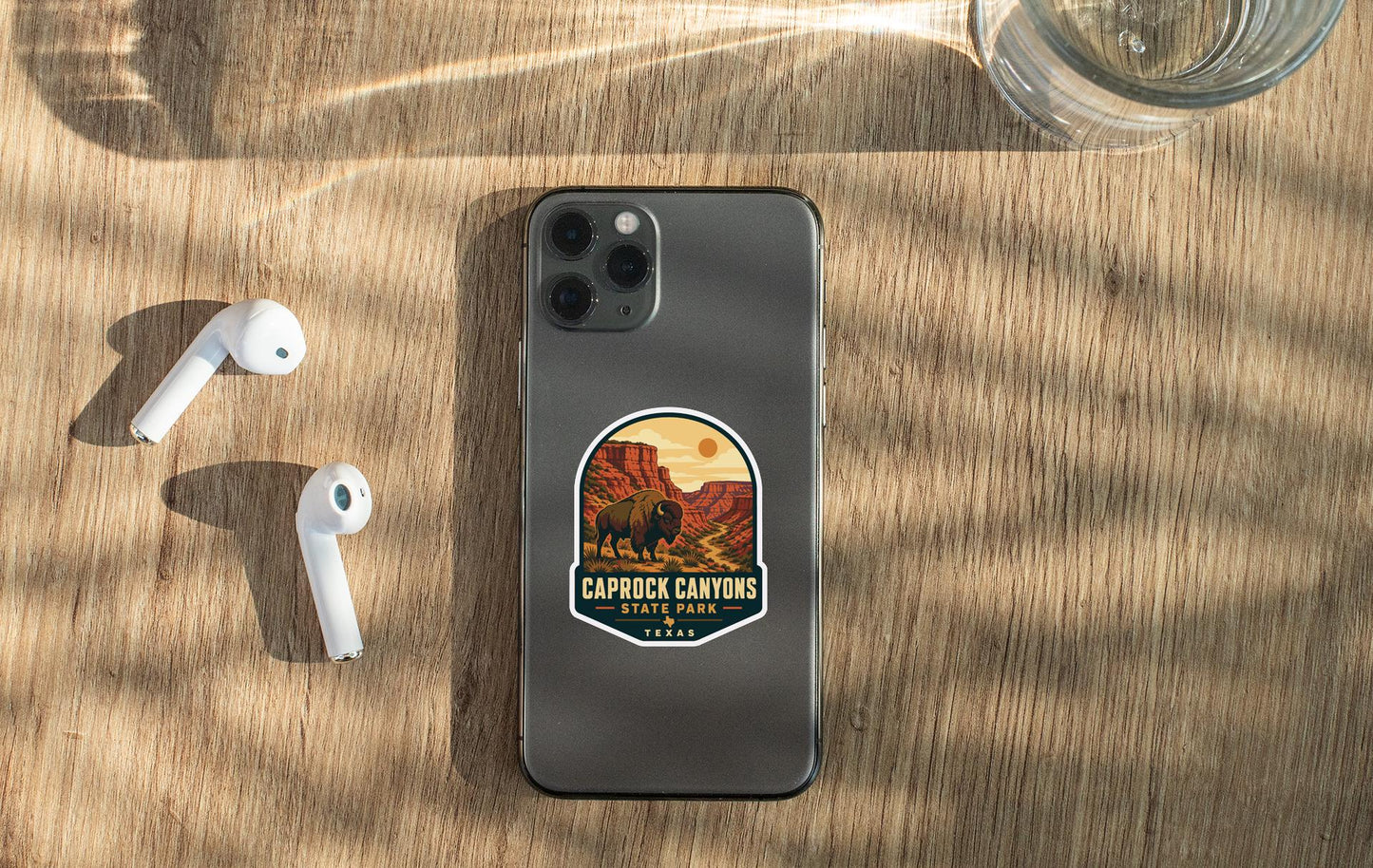 a smartphone with a sticker of the Caprock Canyon National Park, along with a pair of white earbuds and a glass of water on a wooden surface.