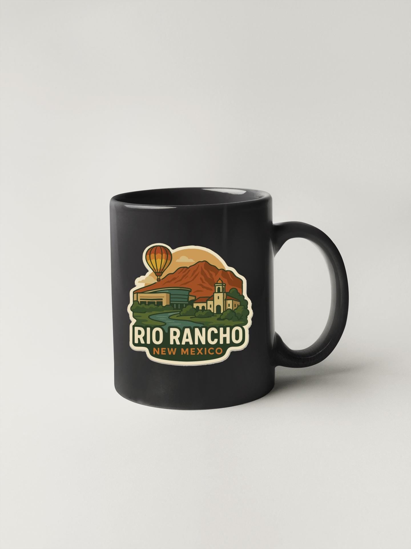 A black coffee mug with a graphic design depicting a hot air balloon, mountains, and a town in New Mexico.