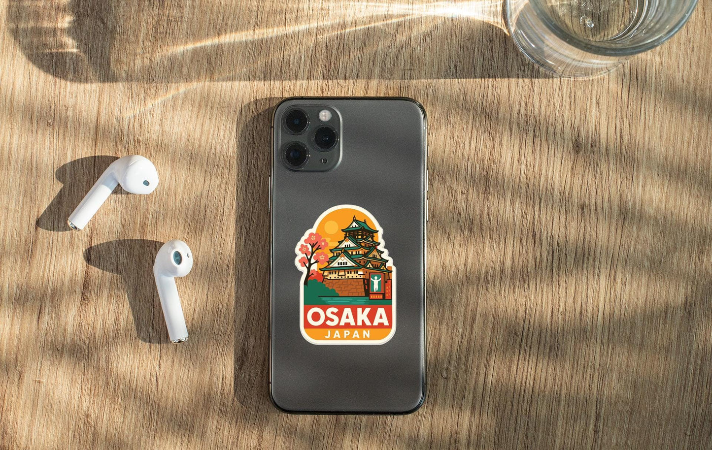 a smartphone with a sticker of Osaka, Japan, placed on a wooden surface alongside a pair of white earbuds and a glass of water.