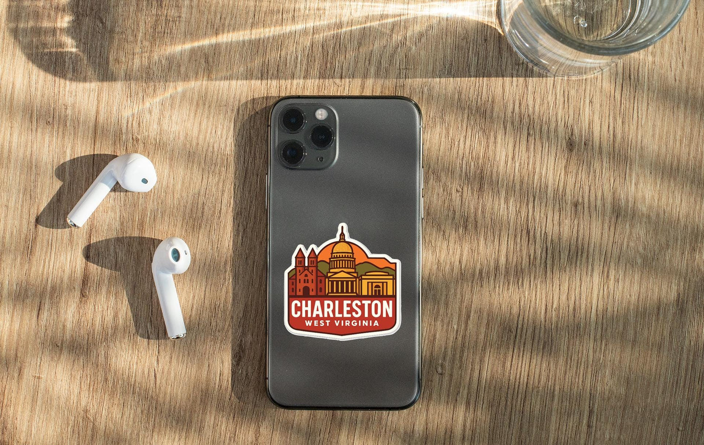 a smartphone with a sticker of Charleston, West Virginia, placed on a wooden surface alongside a pair of white earbuds and a glass of water.