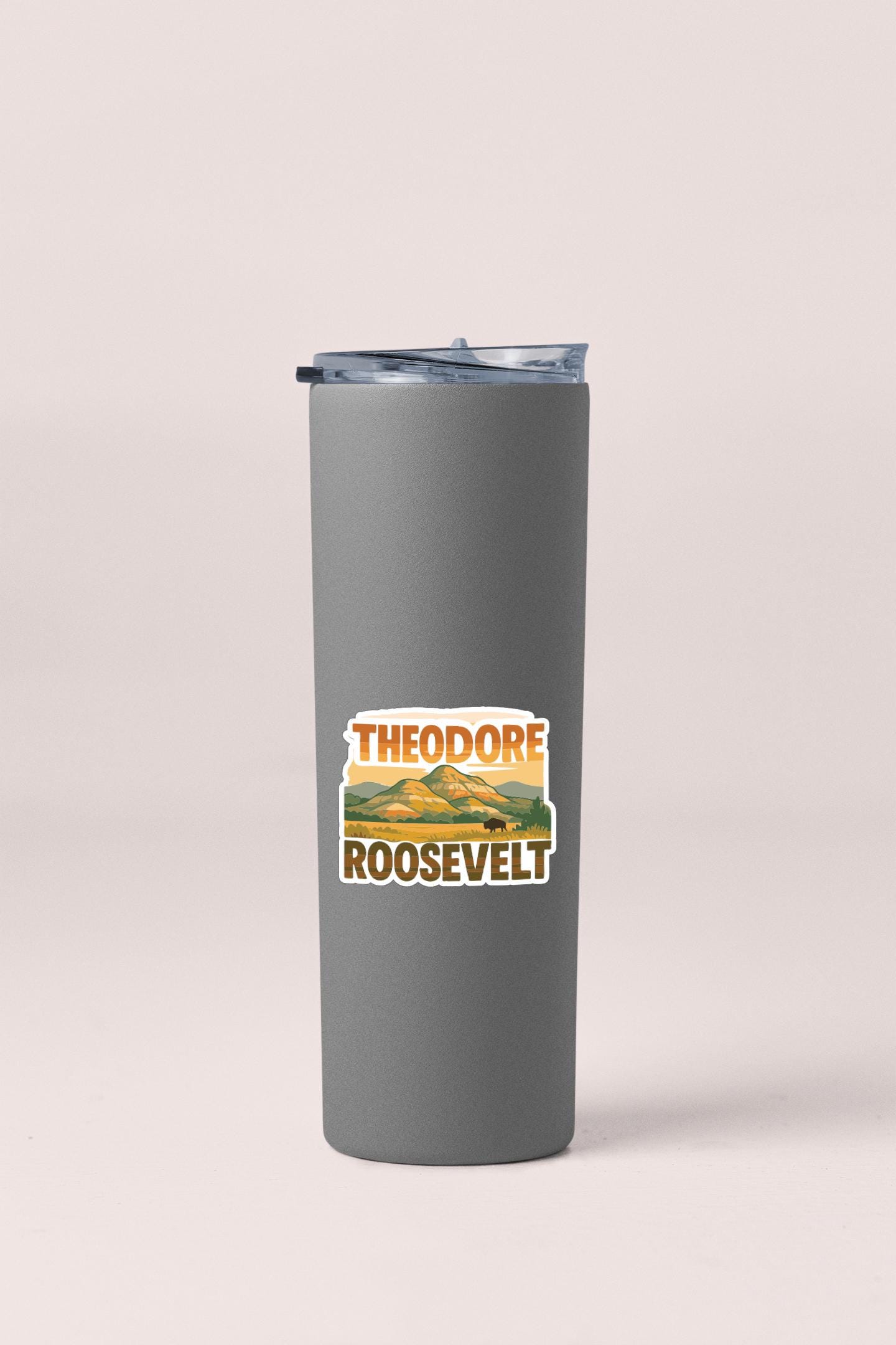 A gray insulated tumbler with a lid and a sticker featuring the text "THEODORE ROOSEVELT" and an illustration of a mountain range.
