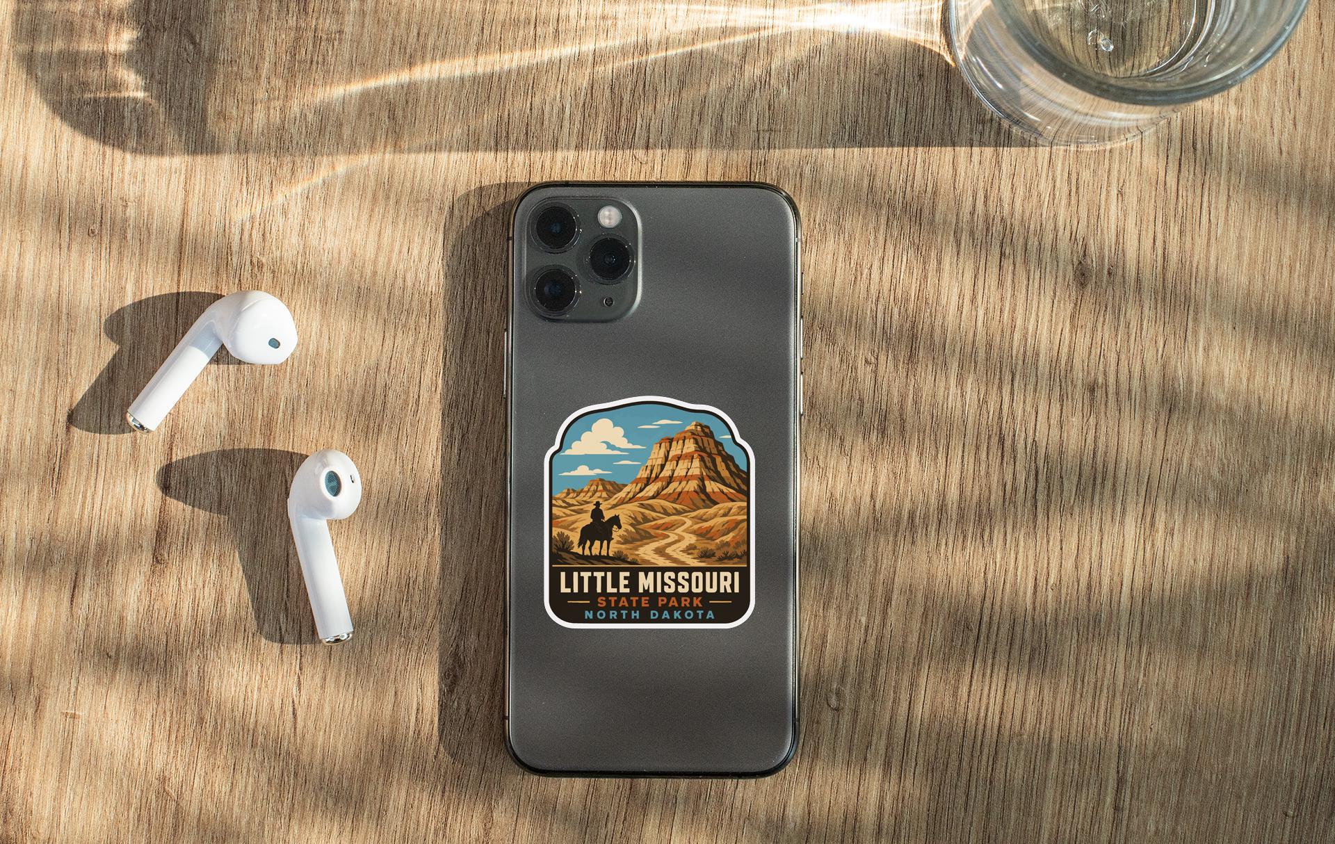 a smartphone with a sticker of Little Missouri State Park on its back, along with a pair of white earbuds and a glass of water on a wooden surface.
