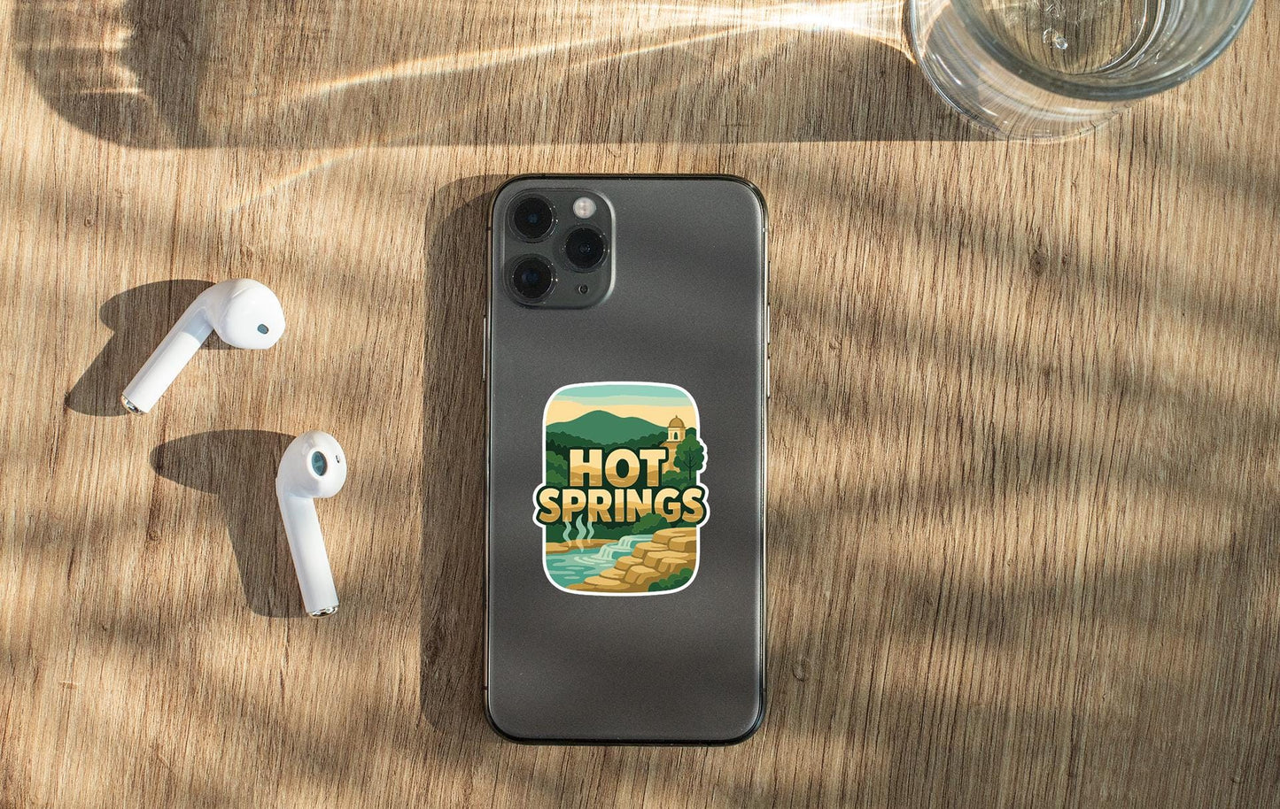 a smartphone with a sticker on the back, placed on a wooden surface alongside a pair of white earbuds and a glass of water.