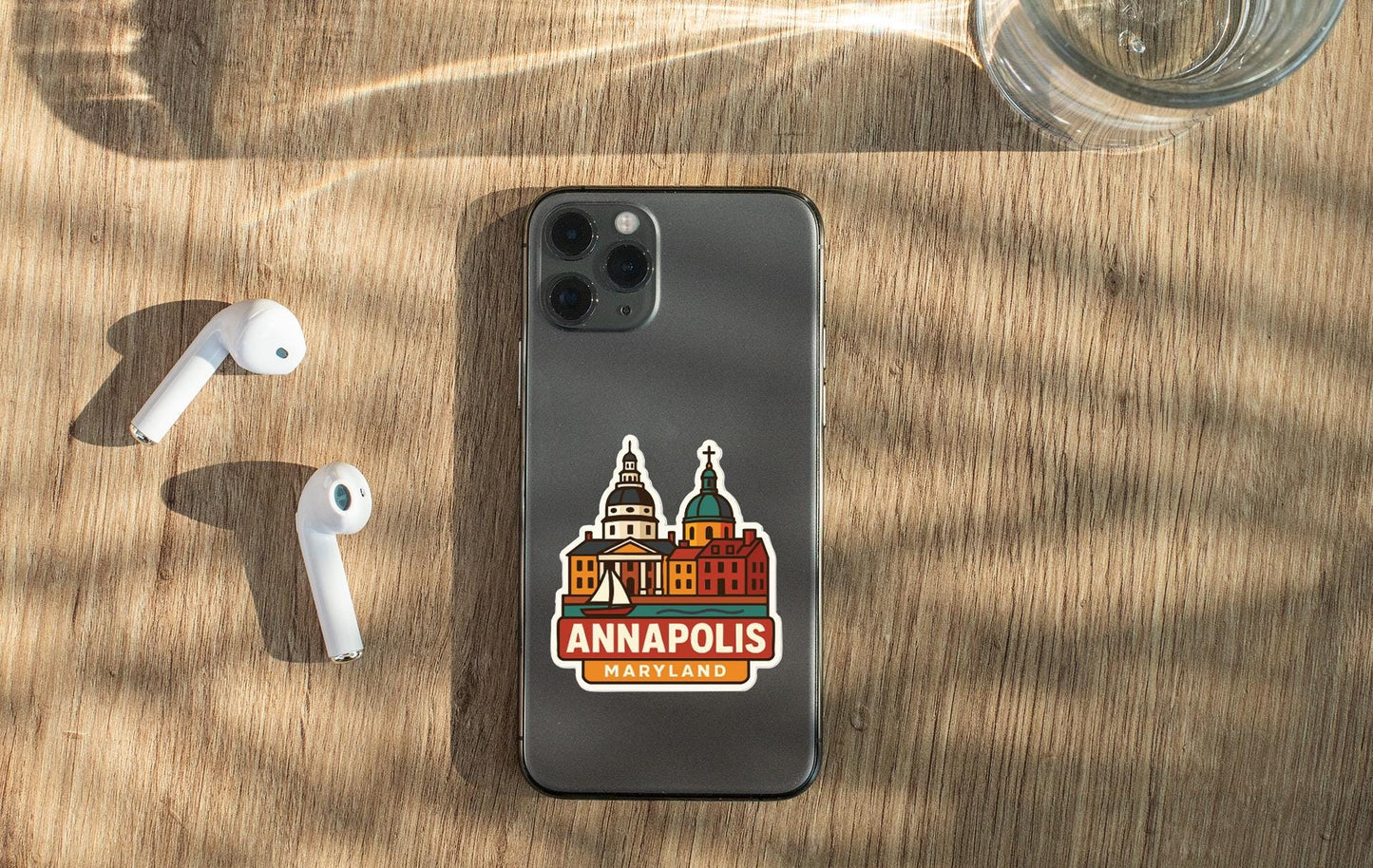 a smartphone with a sticker of Annapolis, Maryland, placed on a wooden surface alongside a pair of white earbuds and a glass of water.