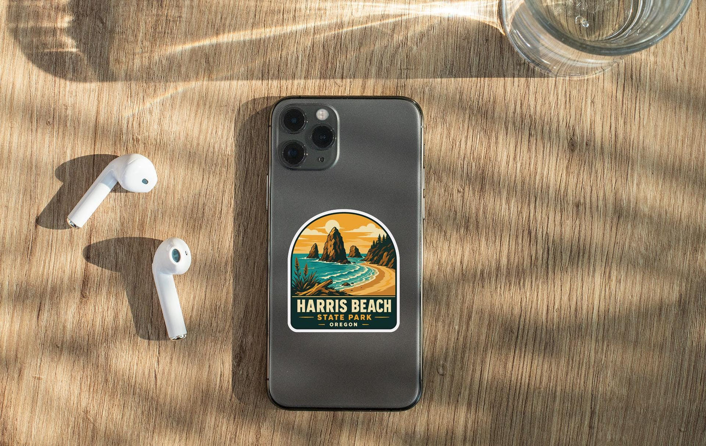 a smartphone with a sticker on the back, placed on a wooden surface alongside a pair of white earbuds and a glass of water.