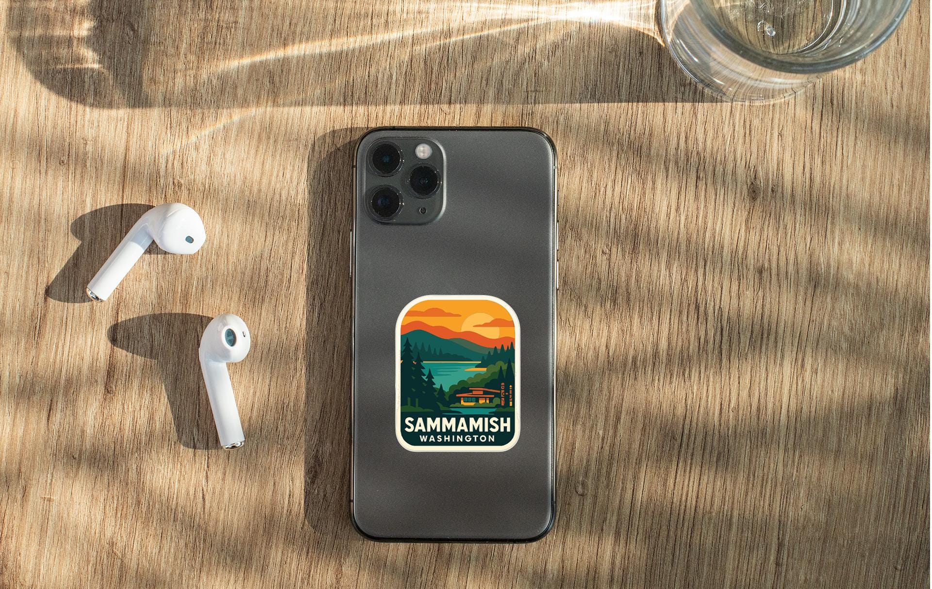 a smartphone with a sticker on the back, placed on a wooden surface alongside a pair of white earbuds and a glass of water.
