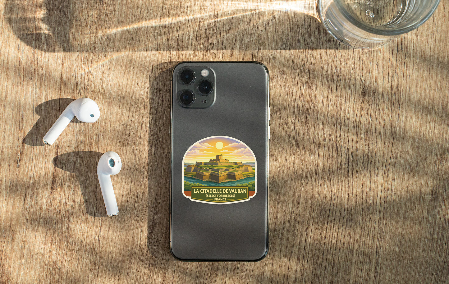 a smartphone with a sticker on the back, placed on a wooden surface alongside a pair of white earbuds and a glass of water.