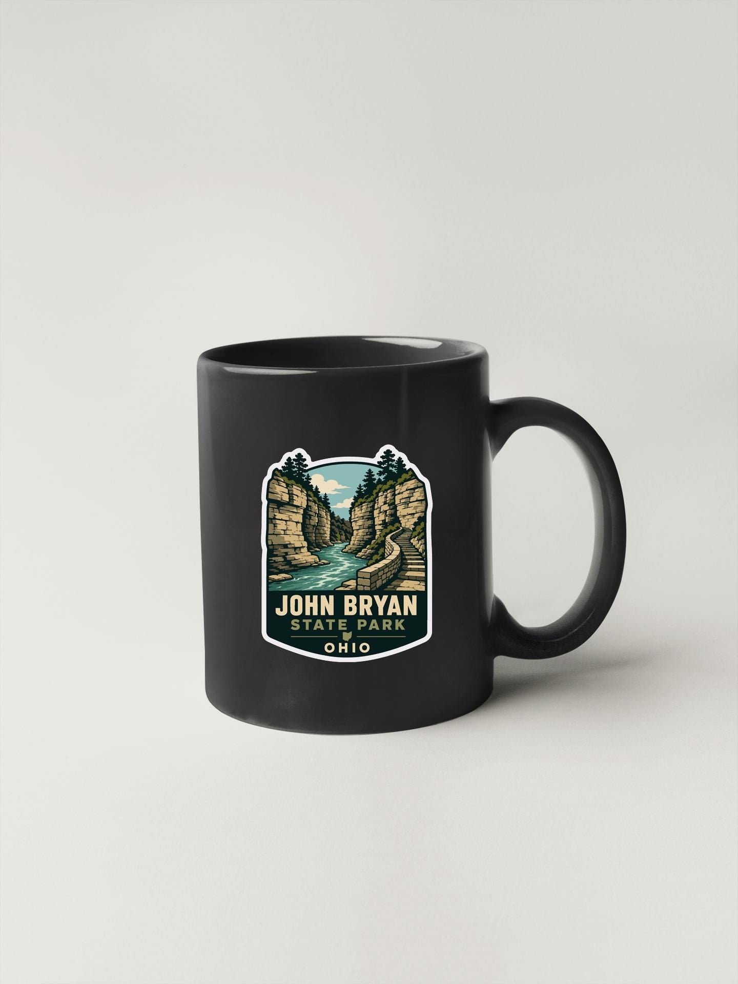 A black coffee mug with a graphic design depicting a scenic landscape featuring a river, cliffs, and trees.