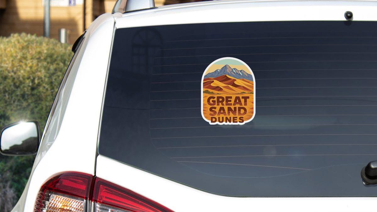 A sticker on the rear window of a white car that says "GREAT SAND DUNES".