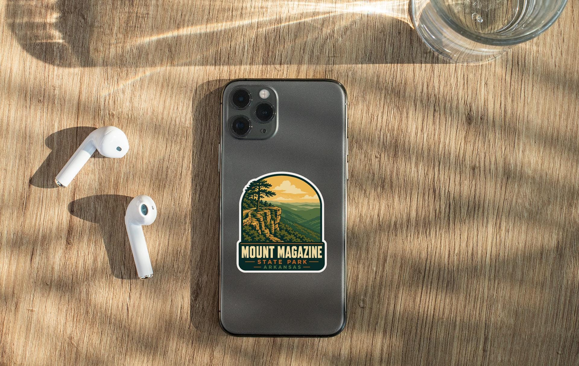 a smartphone with a sticker on it, placed on a wooden surface alongside a pair of white earbuds and a glass of water.