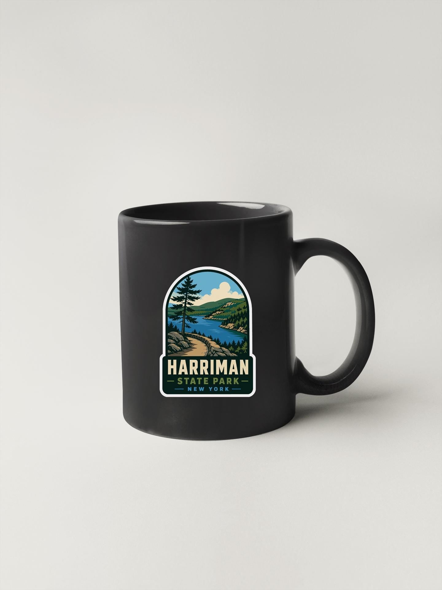 A black coffee mug with a graphic design depicting a scenic landscape, including a river, mountains, and trees.