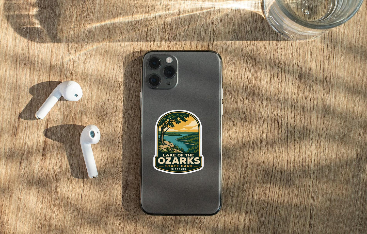 a smartphone with a sticker on the back, placed on a wooden surface alongside a pair of white earbuds and a glass of water.