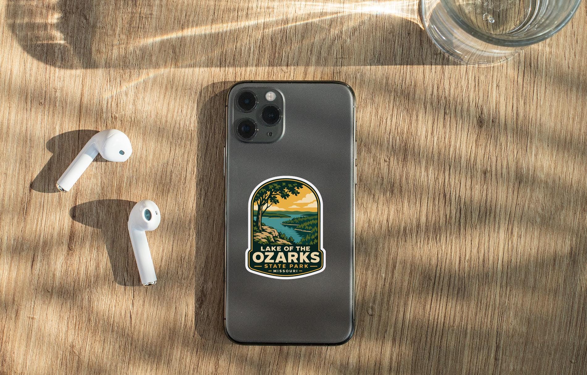a smartphone with a sticker on the back, placed on a wooden surface alongside a pair of white earbuds and a glass of water.