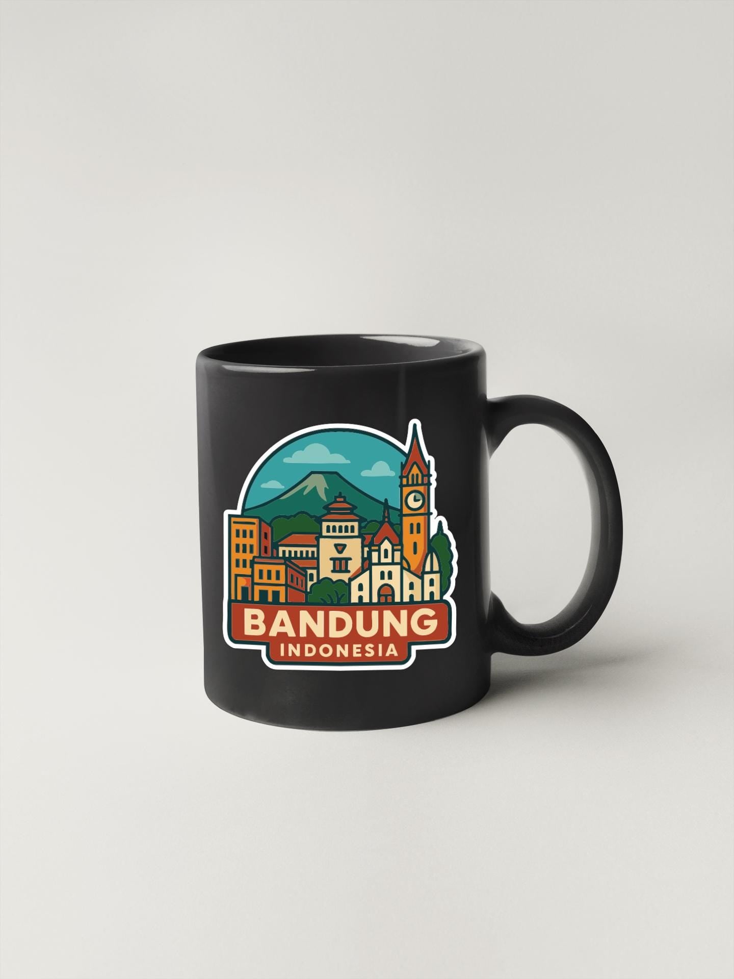 A black coffee mug with a colorful illustration of Bandung, Indonesia, featuring a mountain, buildings, and a clock tower.