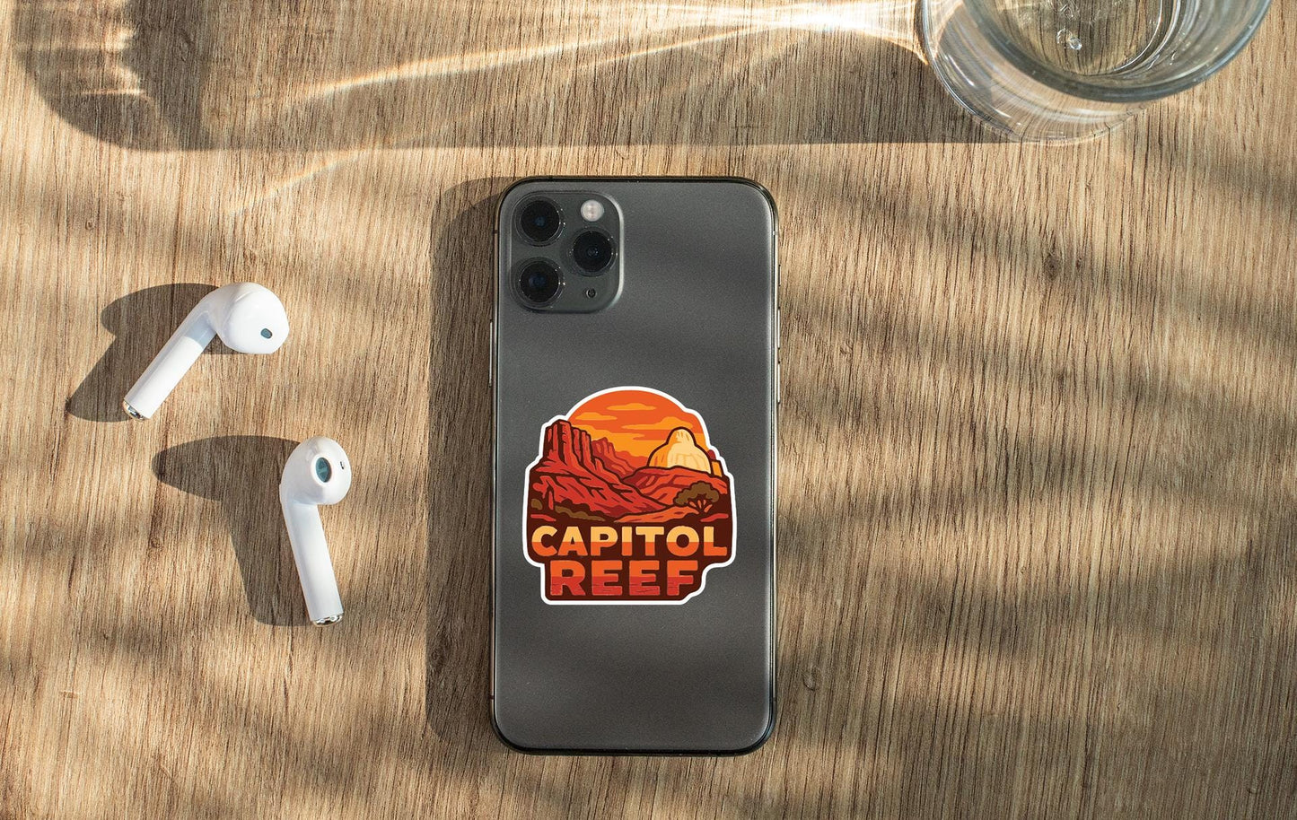 a smartphone with a sticker on it, along with a pair of white earbuds and a glass of water on a wooden surface.