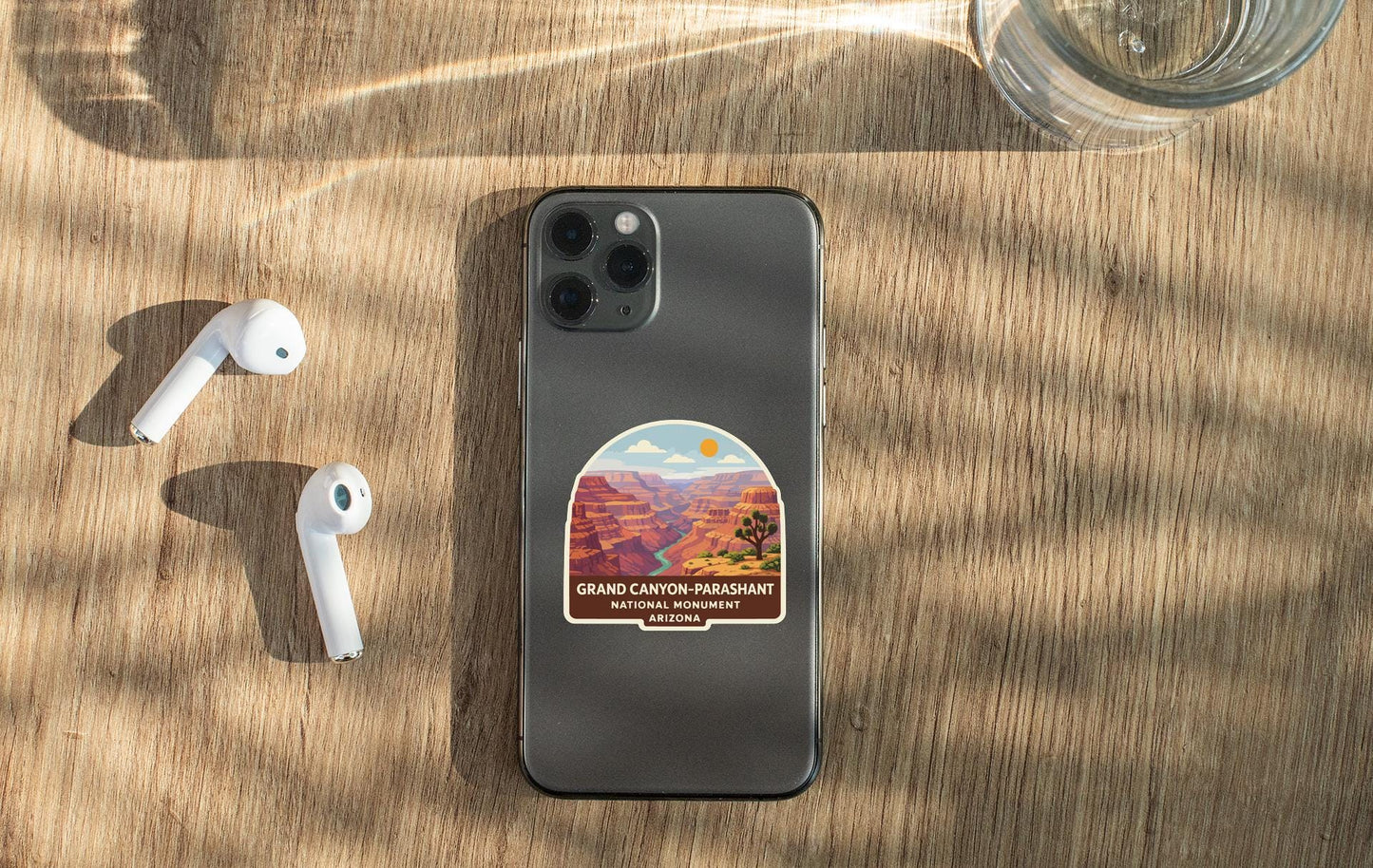 a smartphone with a sticker of the Grand Canyon on it, along with a pair of white earbuds and a glass of water on a wooden surface.