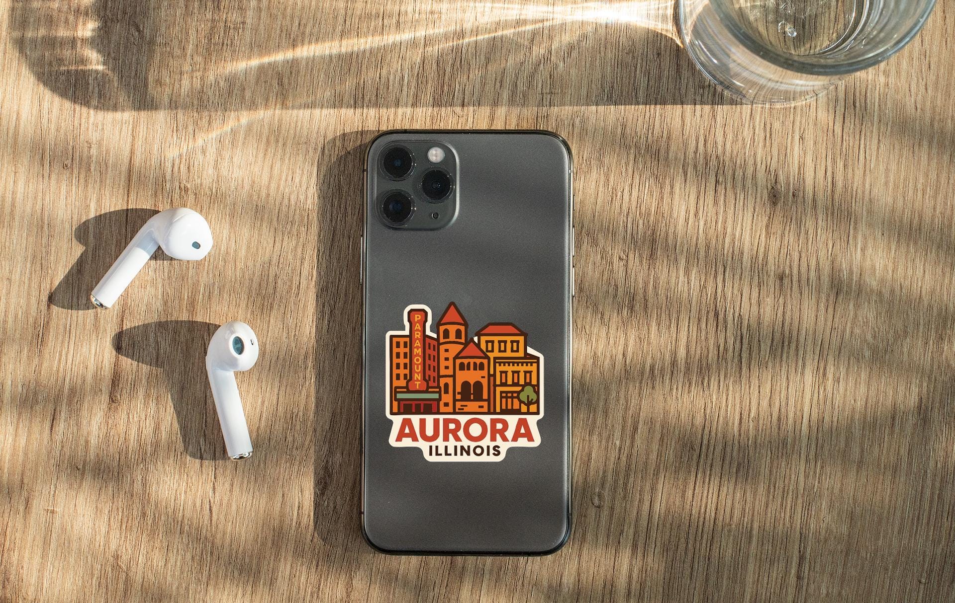 a smartphone with a sticker of a city on it, placed on a wooden surface alongside a pair of white earbuds and a glass of water.