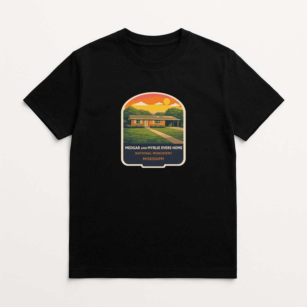 A black t-shirt with a graphic design depicting a house surrounded by trees and a sunset in the background.