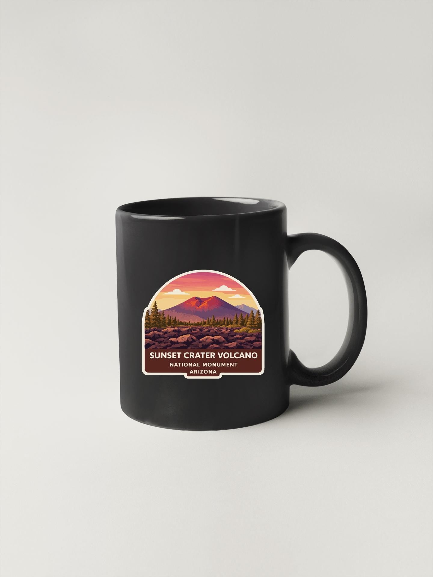A black coffee mug with a graphic design depicting a sunset over a mountain range, with the text "SUNSET CRATER VOLCANO NATIONAL MONUMENT ARIZONA" printed on it.