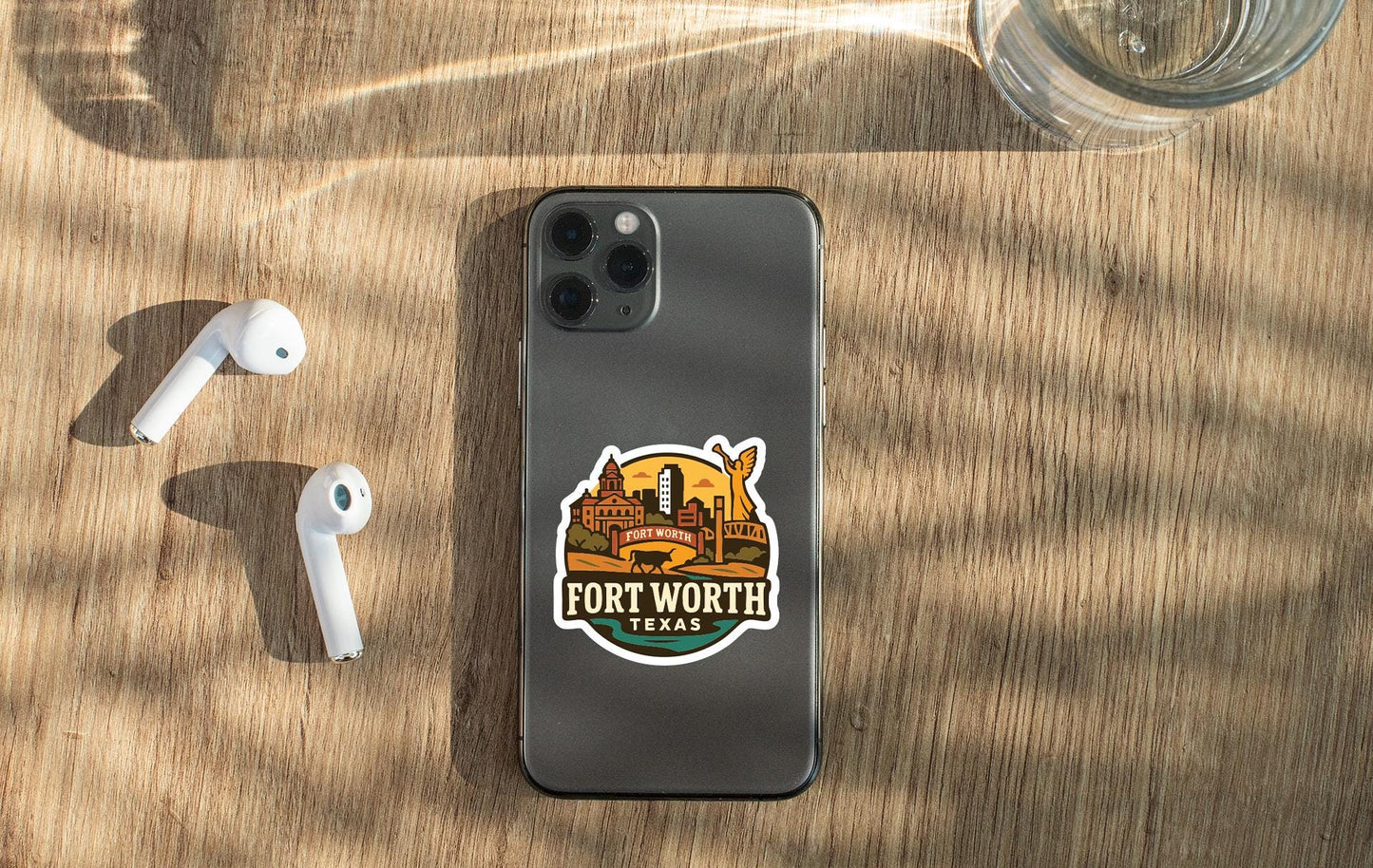 a smartphone with a sticker on the back, placed on a wooden surface alongside a pair of white earbuds and a glass of water.