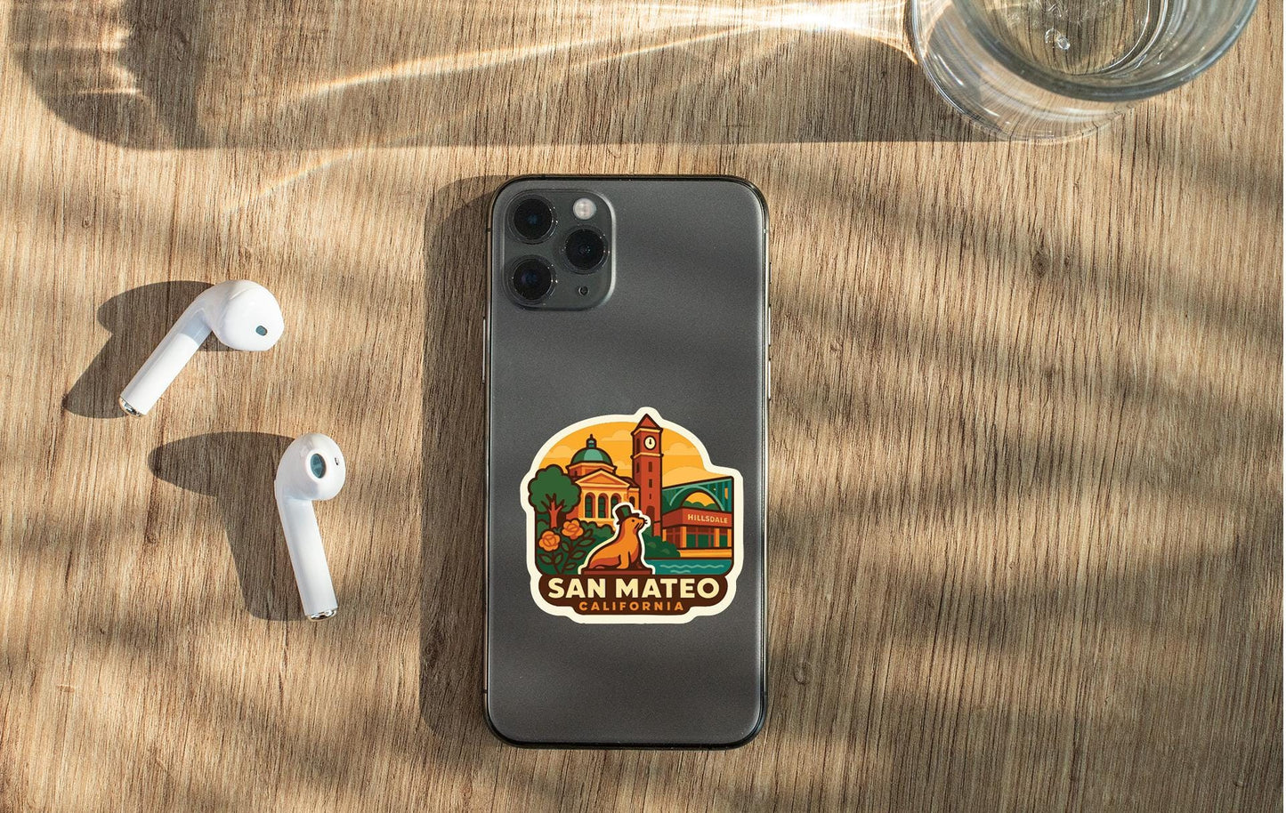 a smartphone with a sticker of San Mateo, California, placed on a wooden surface alongside a pair of white earbuds and a glass of water.