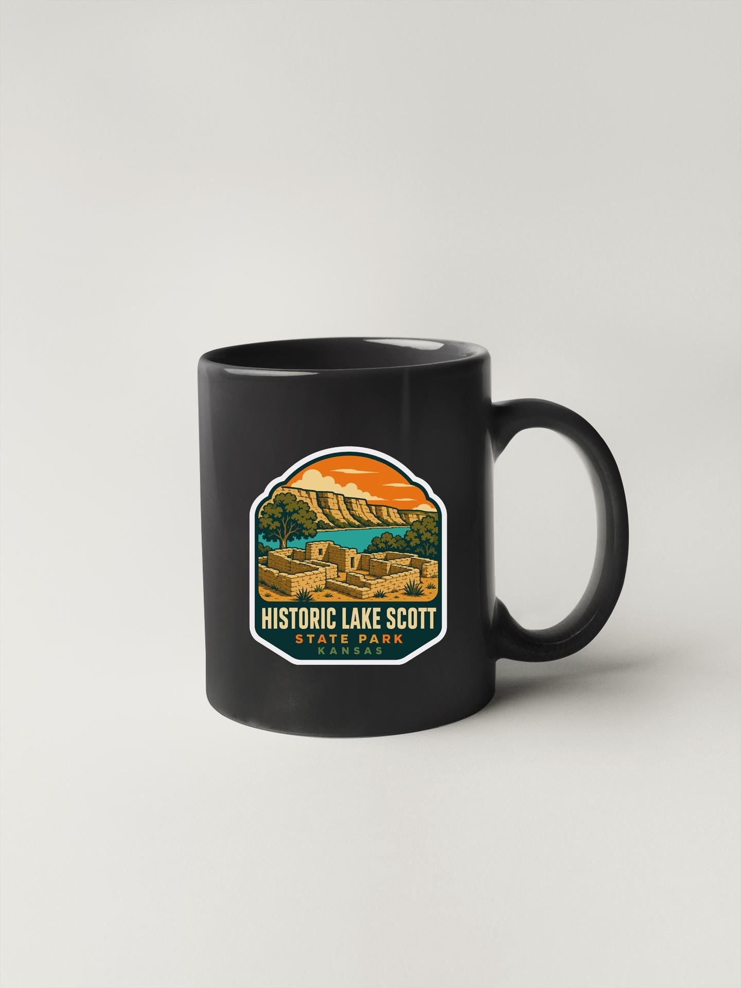A black coffee mug with a colorful graphic depicting a scenic landscape, including mountains, buildings, and a body of water.