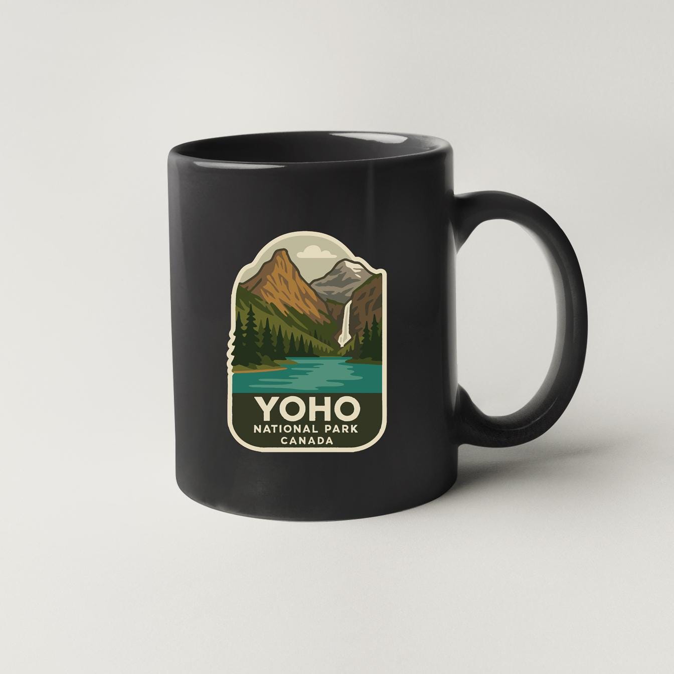A black coffee mug with a graphic design depicting a scenic mountain landscape with a waterfall, representing the Yohoo National Park in Canada.