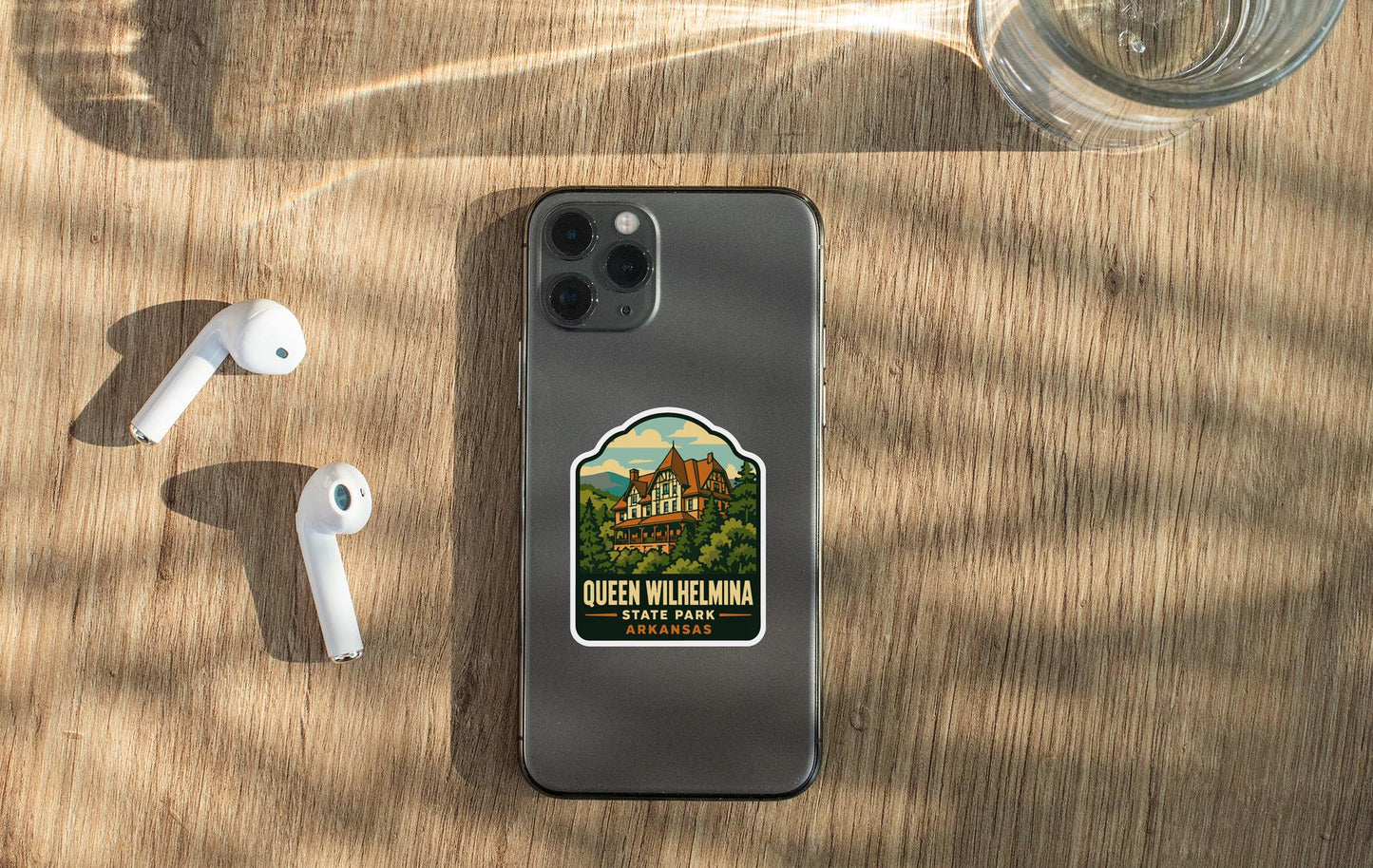 a smartphone with a sticker on it, placed on a wooden surface alongside a pair of white earbuds and a glass of water.