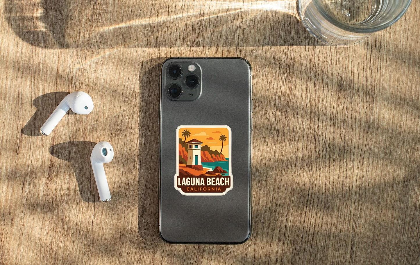 a smartphone with a sticker of Laguna Beach, California, placed on a wooden surface alongside a pair of white earbuds and a glass of water.