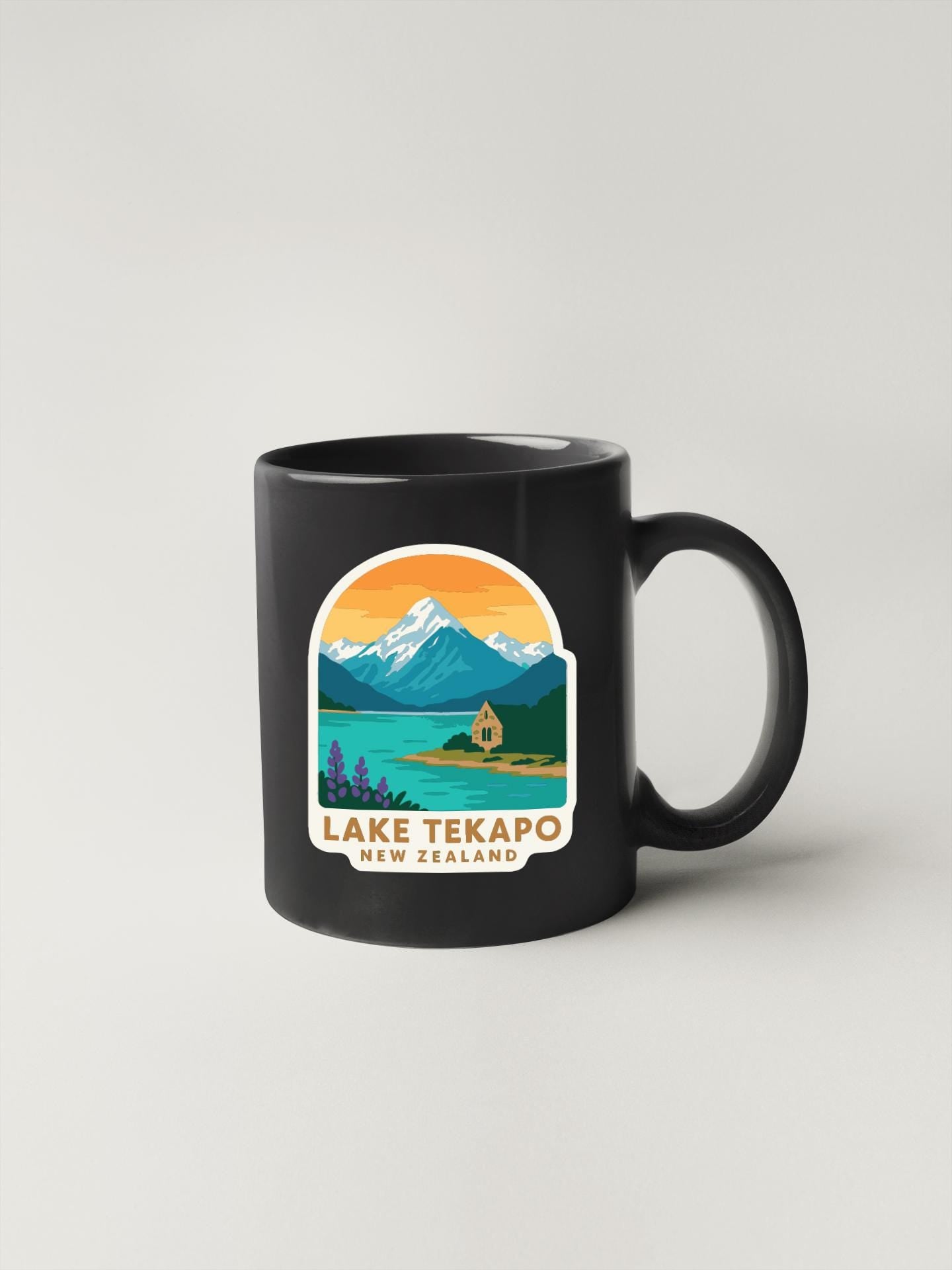 A black coffee mug with a sticker depicting a scenic landscape of Lake Tekapo, New Zealand, featuring mountains, a lake, and a small building.