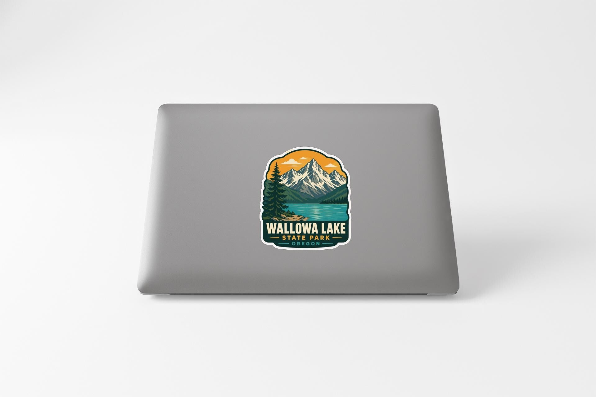 A sticker on the back of a laptop that features a mountain landscape with a lake and pine trees.
