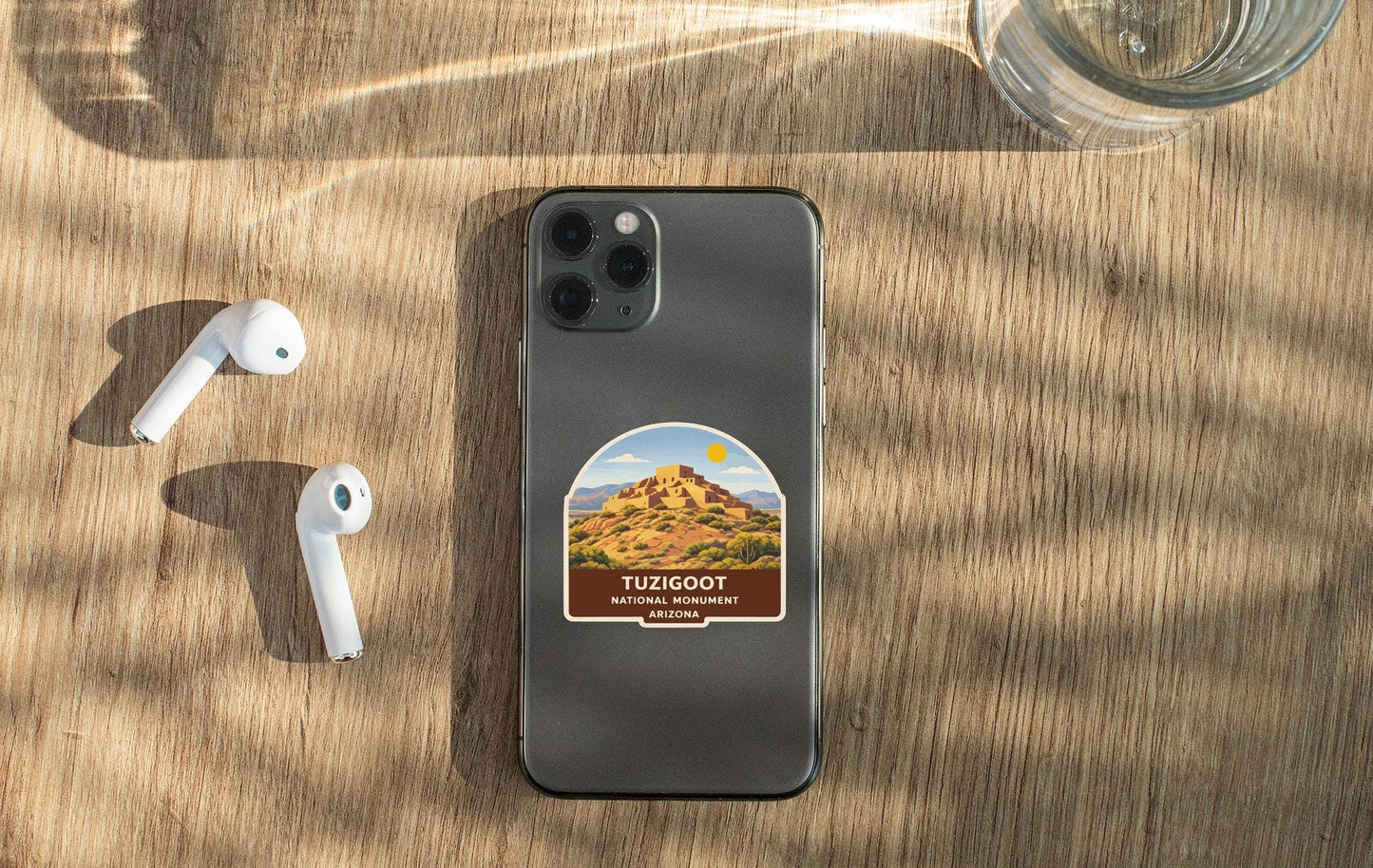 a smartphone with a sticker of a desert landscape on it, placed on a wooden surface alongside a pair of white earbuds and a glass of water.