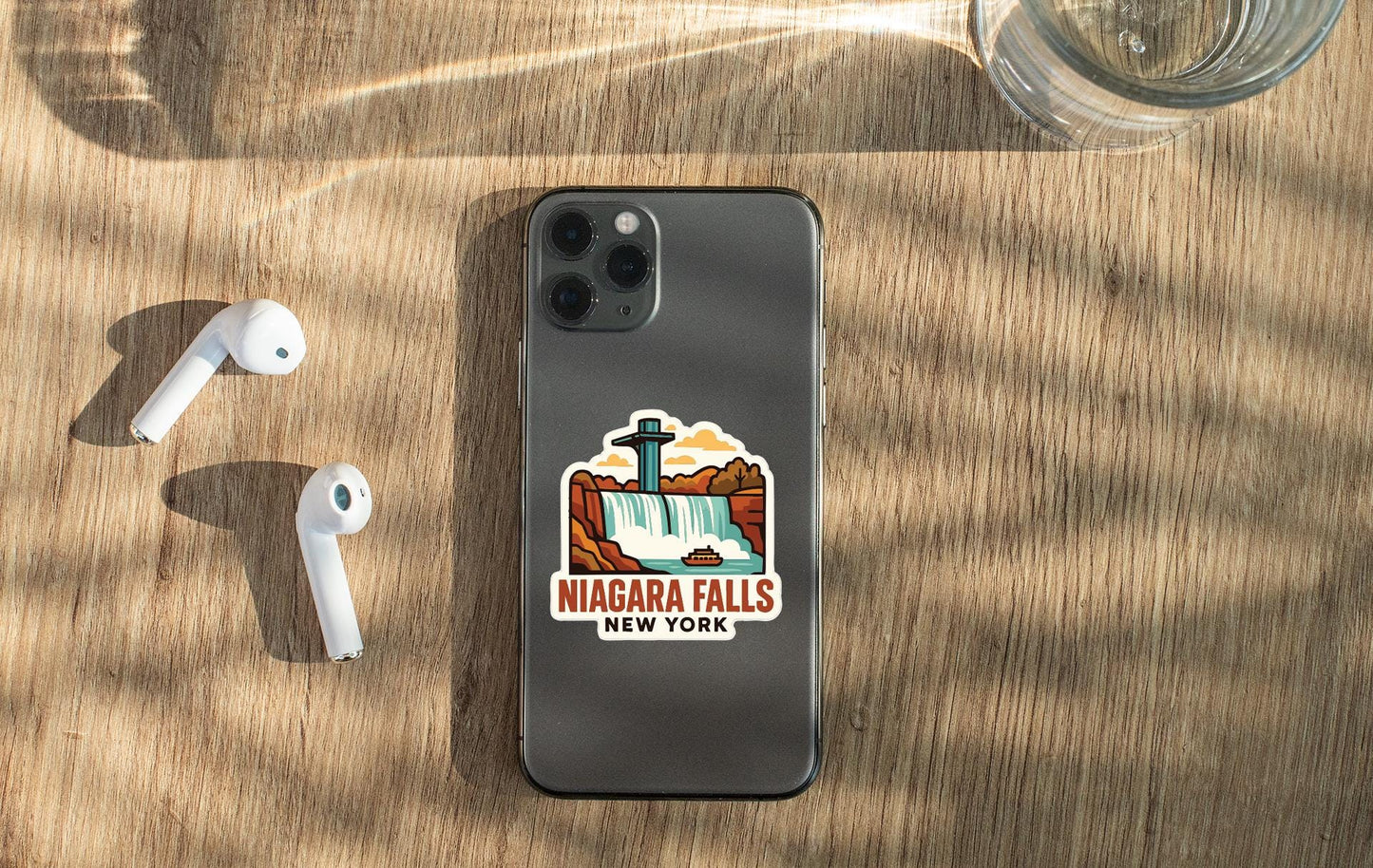 a smartphone with a sticker of Niagara Falls on it, along with a pair of white earbuds and a glass of water on a wooden surface.