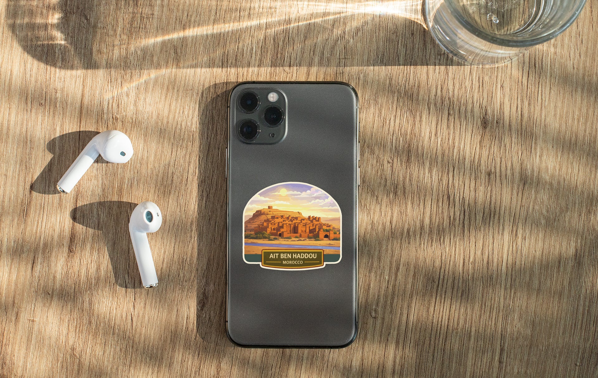 a smartphone with a sticker of a desert landscape on its back, along with a pair of white earbuds and a glass of water on a wooden surface.