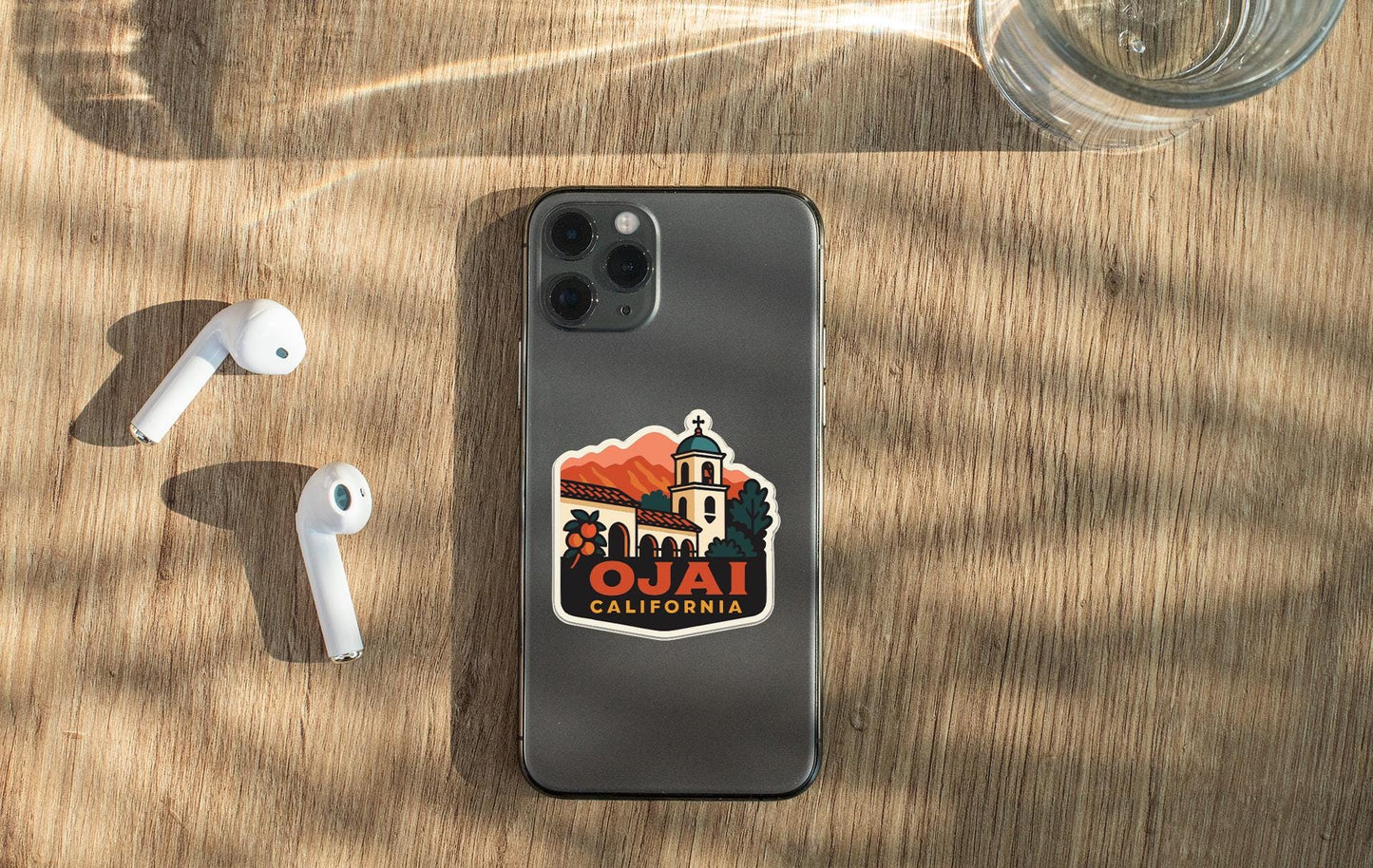 a smartphone with a sticker on the back, placed on a wooden surface alongside a pair of white earbuds and a glass of water.